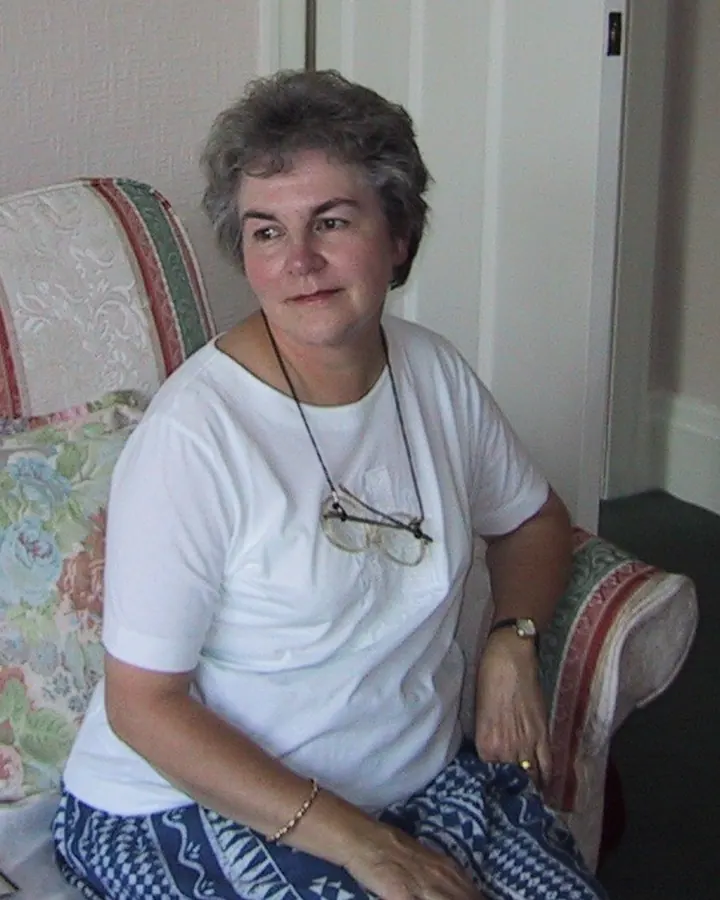 Femme âgée aux cheveux gris portant un haut blanc et un short bleu, assise sur un canapé
