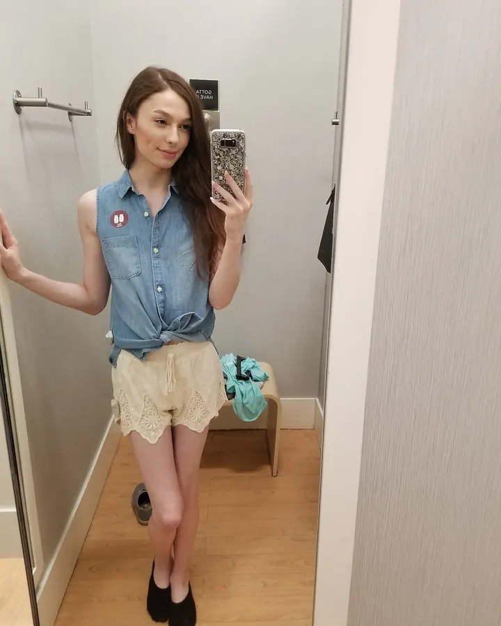 Femme latine aux cheveux châtains portant un haut bleu et un short beige, prenant un selfie dans un miroir