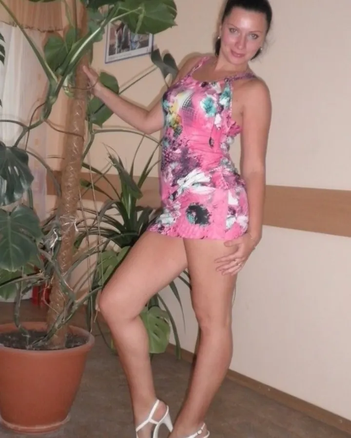 Femme aux cheveux bruns-noirs, yeux bleus, en robe rose imprimée florale et talons blancs, posant près d'une plante verte.
