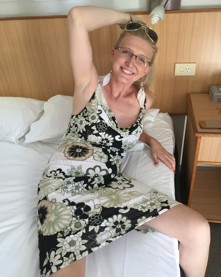 Femme blonde aux cheveux mi-longs, portant une robe à motifs floraux, assise sur un lit dans une chambre d'hôtel.