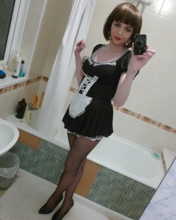 Femme en robe noire avec lacets blancs, collants résille, prenant un selfie dans une salle de bains