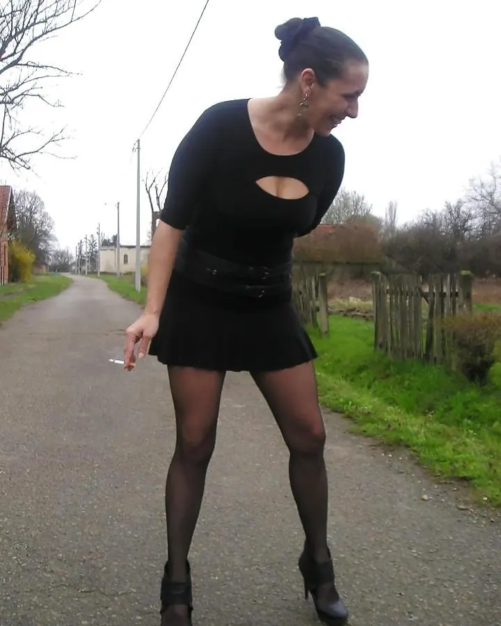 Femme aux cheveux foncés, vêtue d'une robe noire à découpes, tenant une cigarette, debout sur une route rurale.