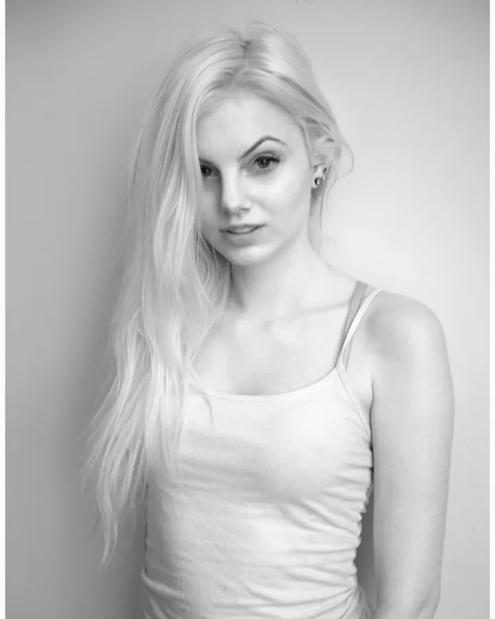 Femme blonde, en haut noir et jeans gris, posant devant un miroir, souriant à la caméra.