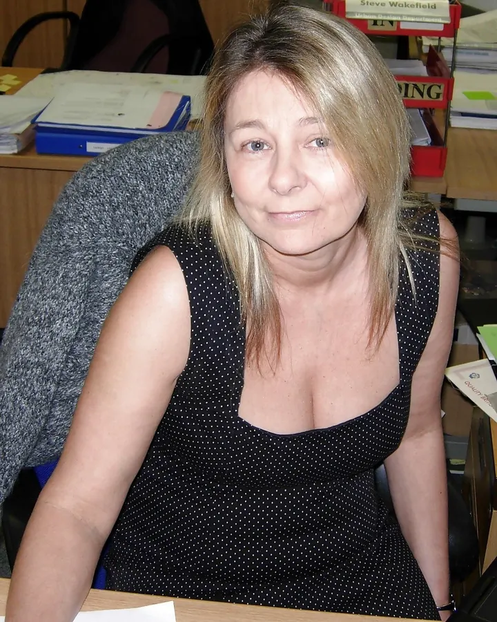 Femme blonde, assise à un bureau, vêtue d'une robe noire à pois, regardant la caméra avec un léger sourire.