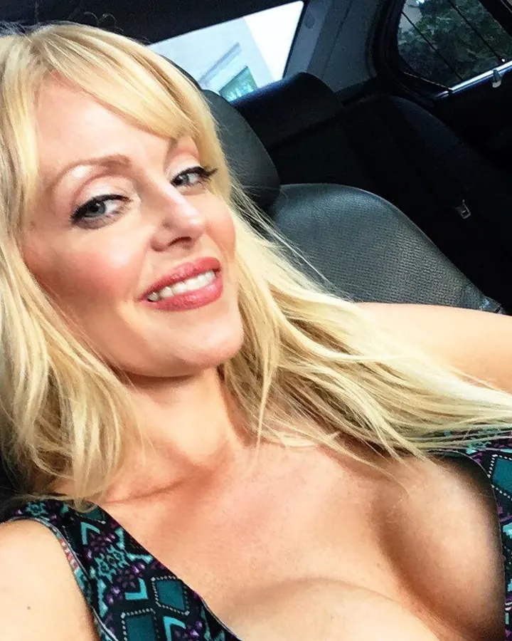 Femme blonde, assise dans une voiture, souriant à la caméra, portant un haut à motifs colorés.