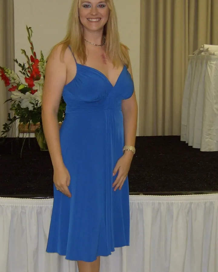 Femme blonde souriante, debout, portant une robe bleue ajustée, avec des sandales à talons, devant une table décorée de fleurs.
