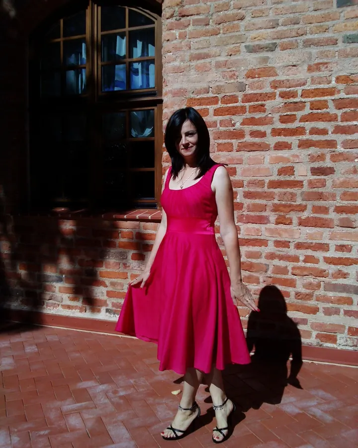 Femme aux cheveux bruns-noirs portant une robe rose vif, debout devant un mur en briques rouges avec une fenêtre en bois.