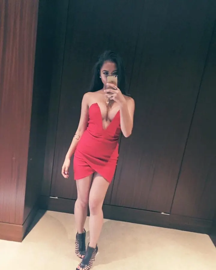 Femme aux cheveux bruns-noirs portant une robe rouge profondément décolletée, prenant un selfie devant un mur sombre.