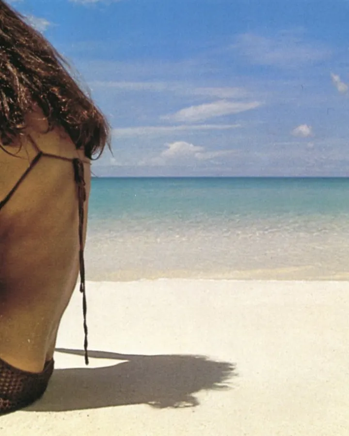 Femme aux cheveux bruns longs, portant un maillot de bain noir à motifs, assise sur le sable d'une plage avec l'océan turquoise et un ciel bleu en arrière-plan.