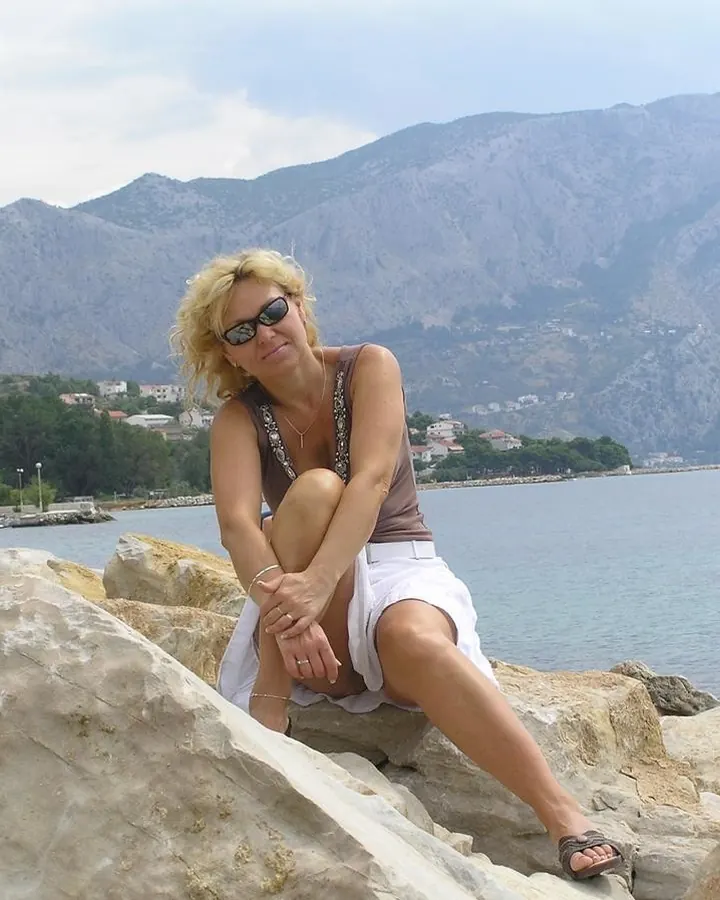 Femme blonde aux cheveux courts, portant un haut beige et une jupe blanche, assise sur des rochers près d'une mer bleue avec des montagnes en arrière-plan.