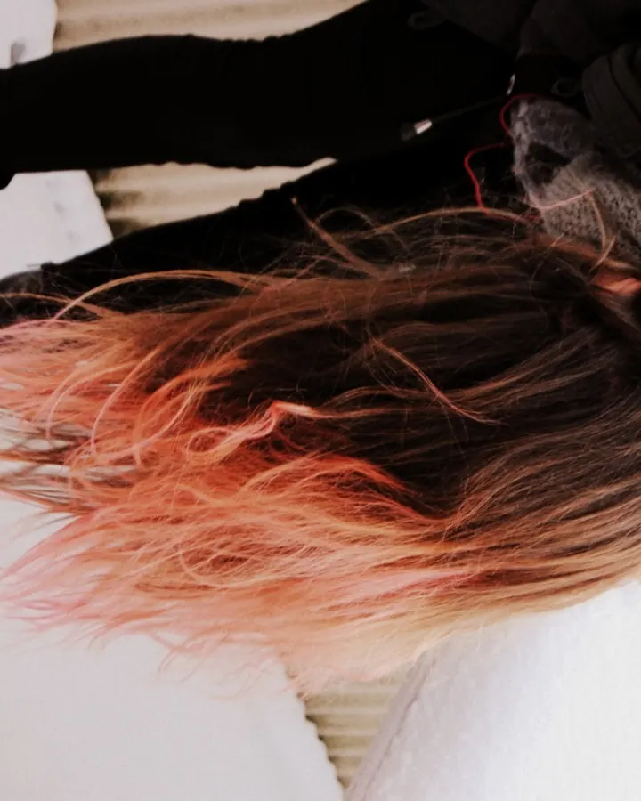 Femme aux cheveux roux, allongée dans la neige, vêtue de noir, avec des cheveux teints en rose au bout.