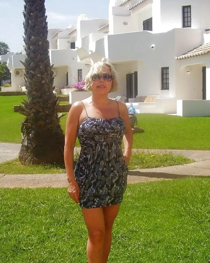 Femme blonde en robe imprimée, debout sur une pelouse verte devant des bâtiments blancs.