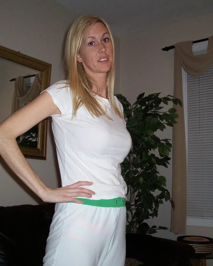 Femme blonde, vêtue d'un t-shirt blanc et d'un pantalon blanc, posant dans un salon.
