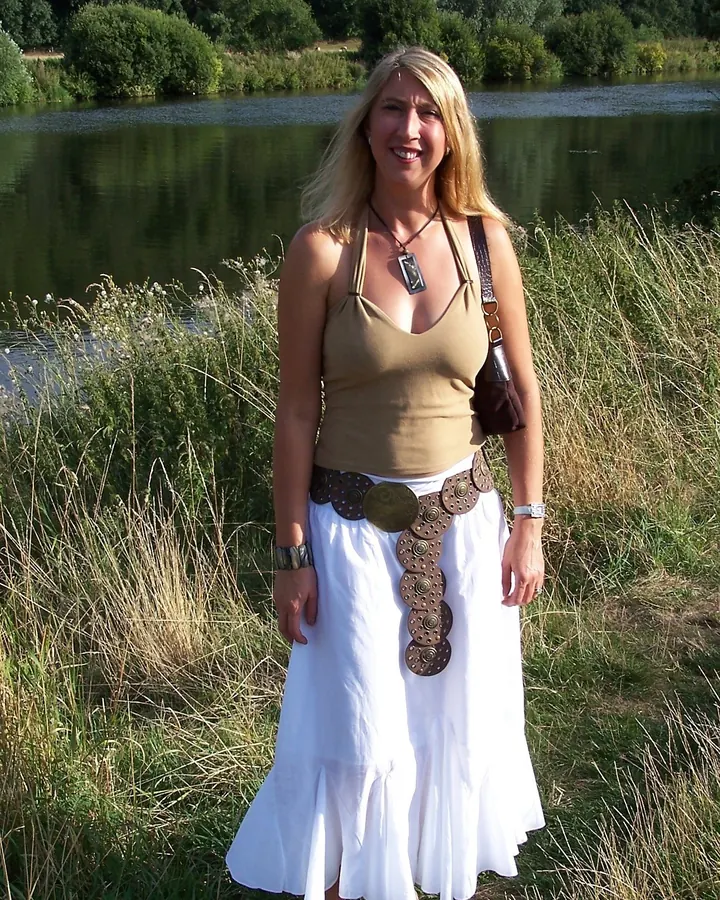 Femme blonde, portant une robe blanche et un haut beige, debout près d'un lac.