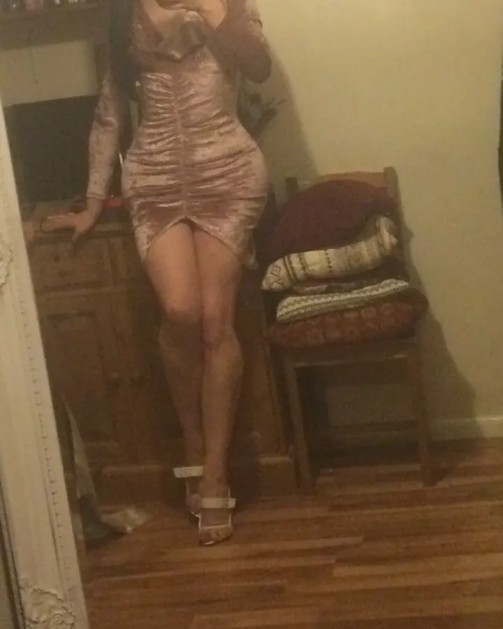 Femme aux cheveux bruns portant une robe moulante dorée, debout devant un miroir dans une pièce éclairée tamisée