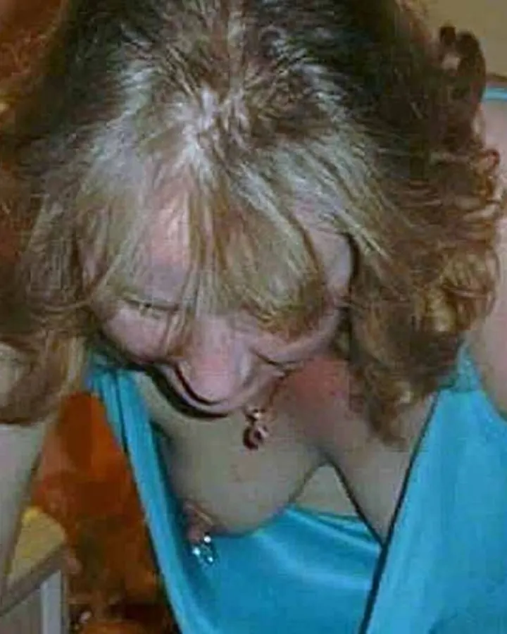 Femme blonde en haut bleu, penchée en avant, riant, dans une pièce éclairée par une lampe