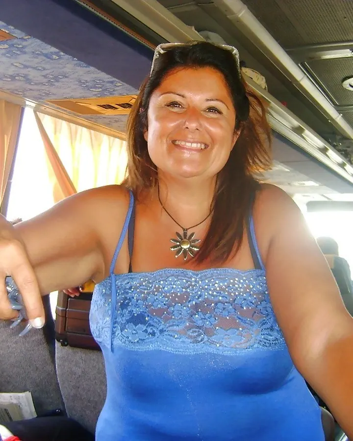 Femme latino souriante, portant un haut bleu à dentelle, assise dans un bus avec des rideaux orange.