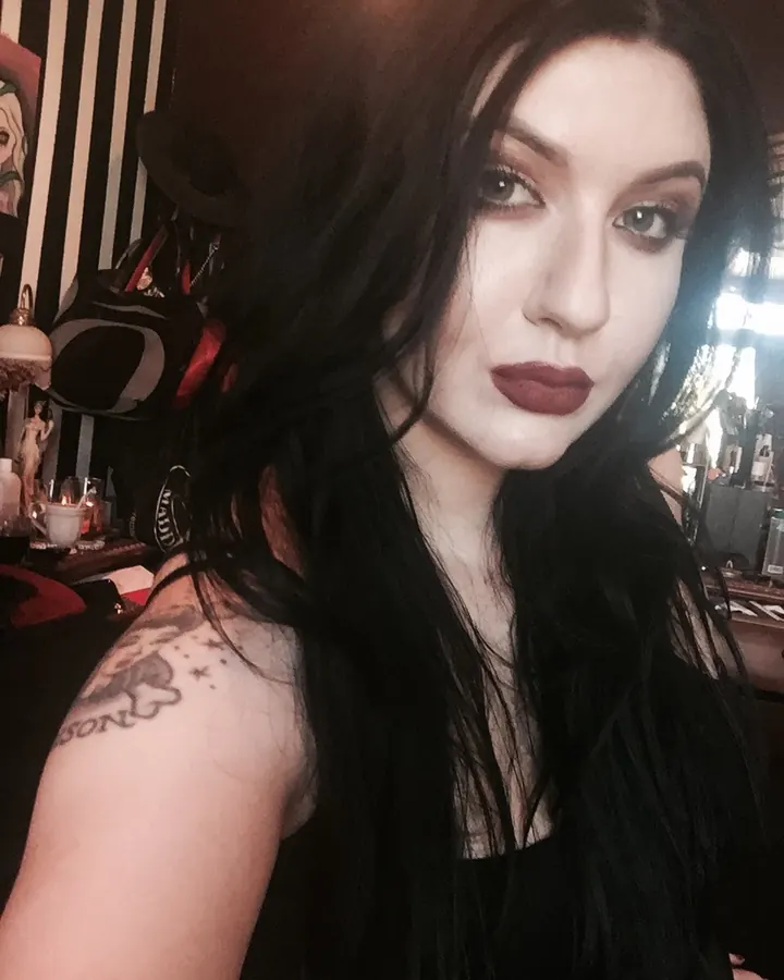 Femme aux cheveux bruns, portant un haut noir à sequins, avec un collier fin et un tatouage visible sur l'épaule.