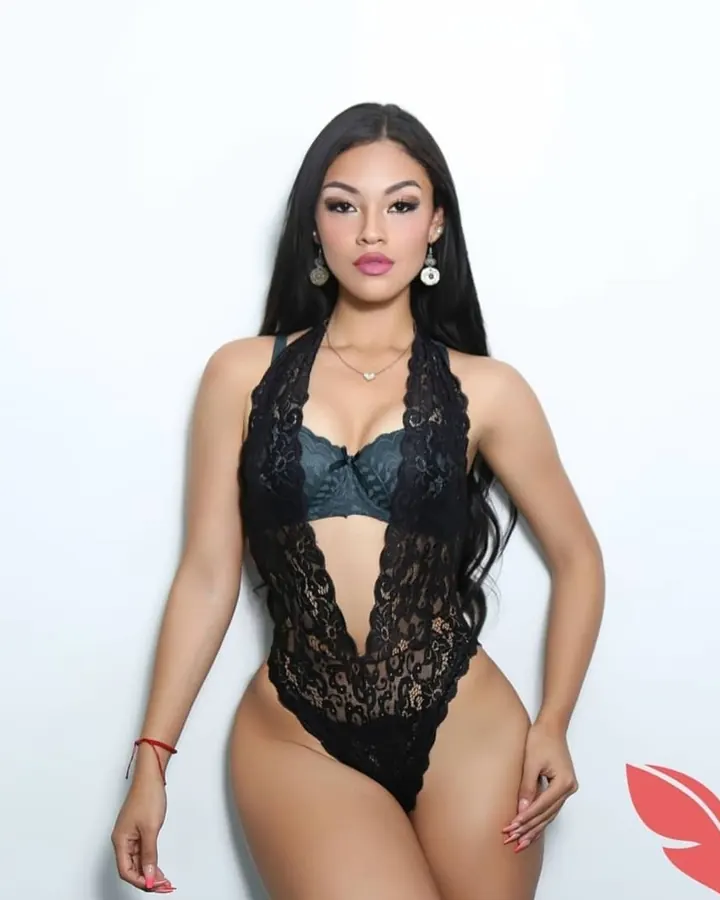 Femme latina souriante, portant un haut noir à dentelle, posant avec un regard direct et un sourire charmeur.