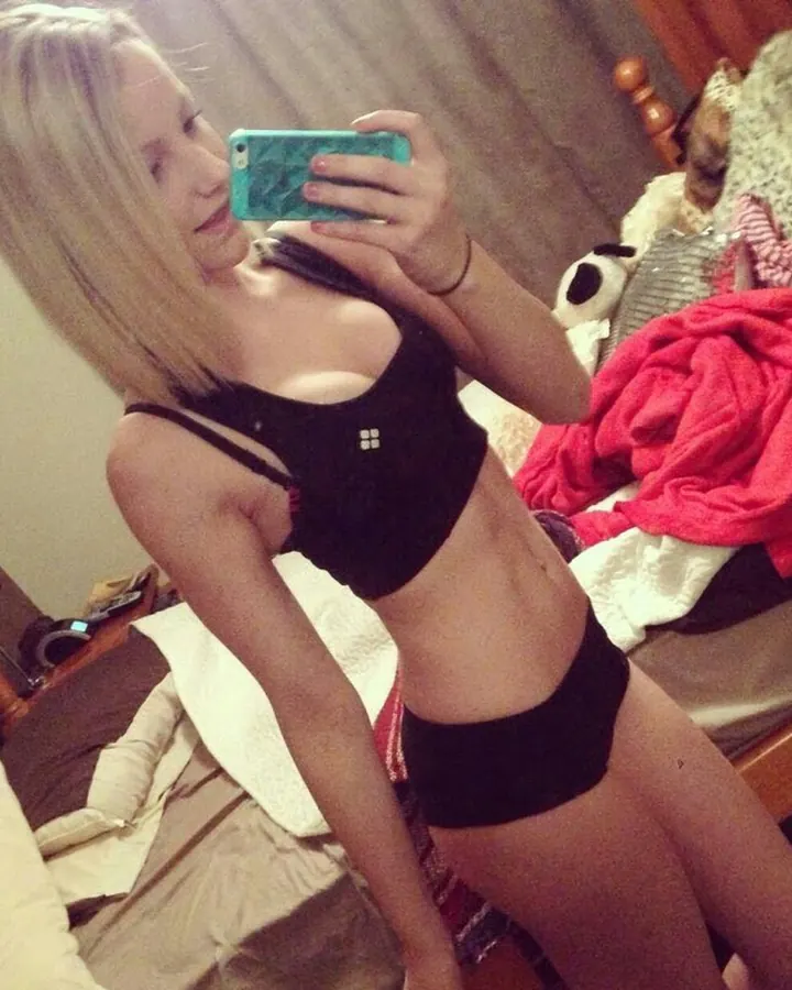 Jeune femme blonde en sous-vêtements noirs, prenant un selfie devant un lit avec des couvertures colorées.