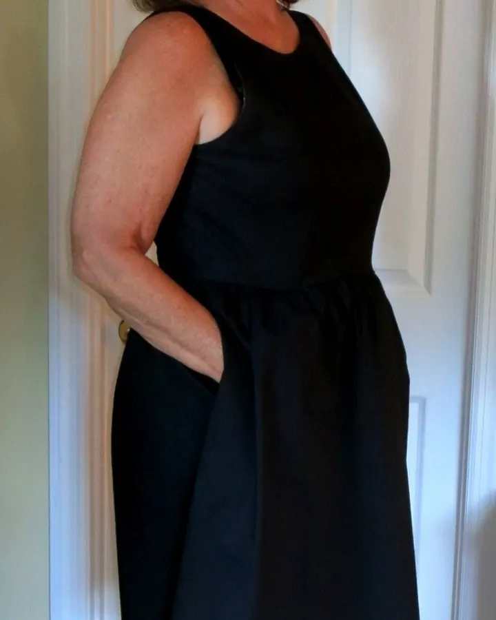 Femme de 45 à 60 ans en robe noire, lunettes, souriante, pieds en talons argentés devant une porte blanche.