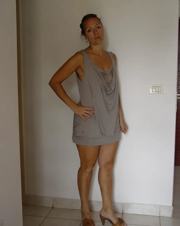 Femme en robe grise, main sur la hanche, debout près d'un mur blanc, regardant l'appareil.