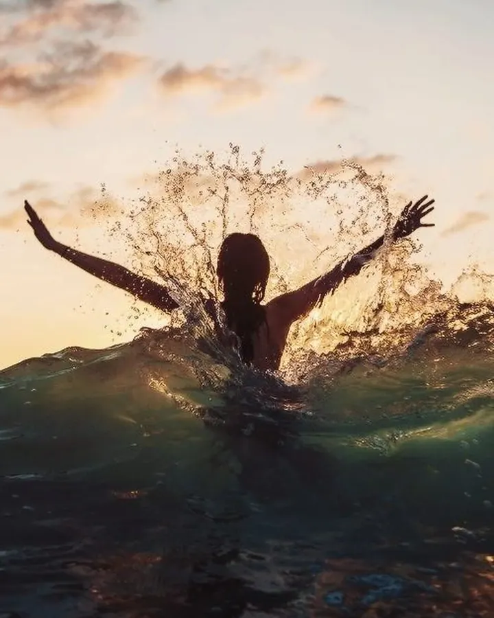 Silhouette féminine dans l’eau, bras écartés, éclaboussures, coucher de soleil en arrière-plan.