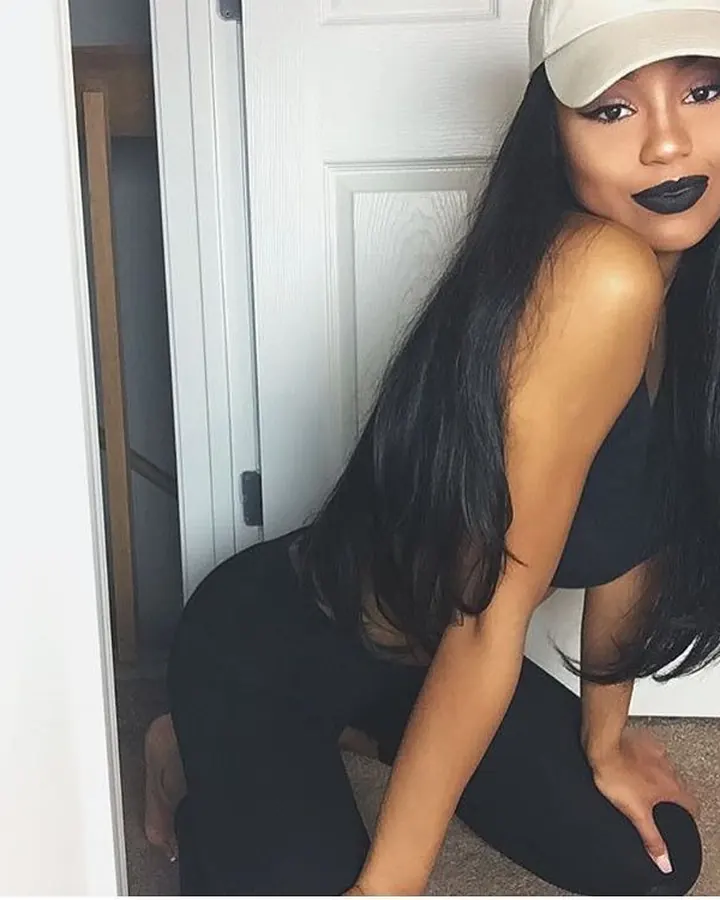 Jeune femme aux longs cheveux noirs, casquette beige, lèvres noires, agenouillée près d'une porte blanche, regardant vers l'appareil.