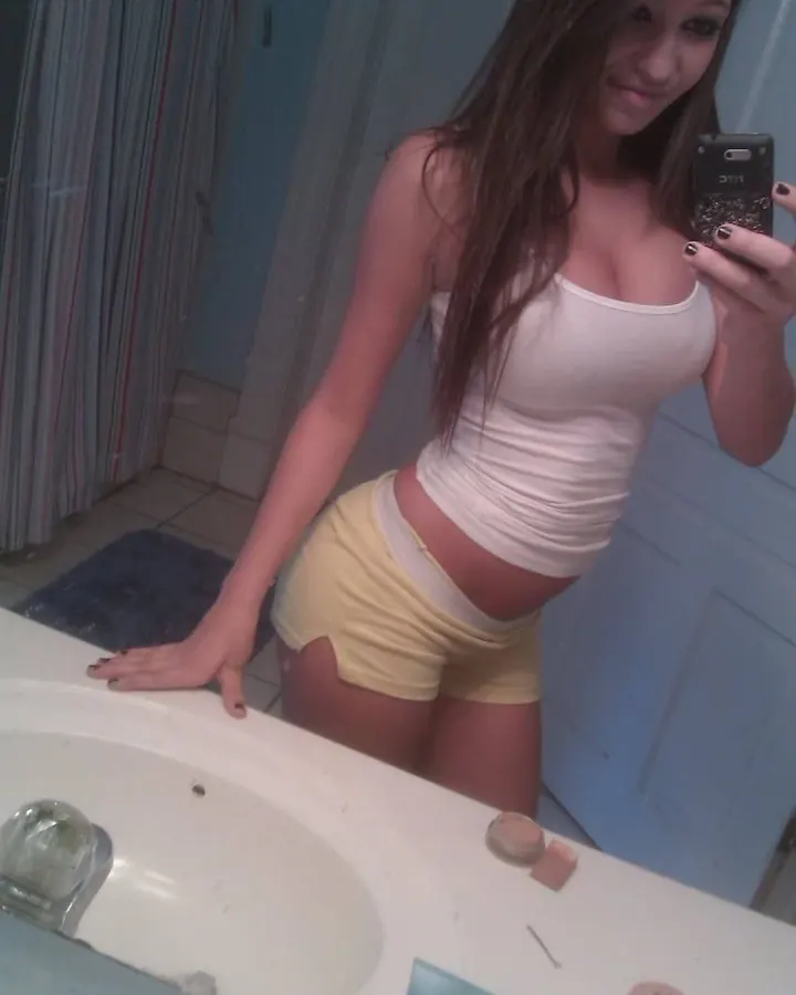 Jeune femme en haut blanc et shorts jaunes, prenant un selfie dans une salle de bains, main sur le lavabo.
