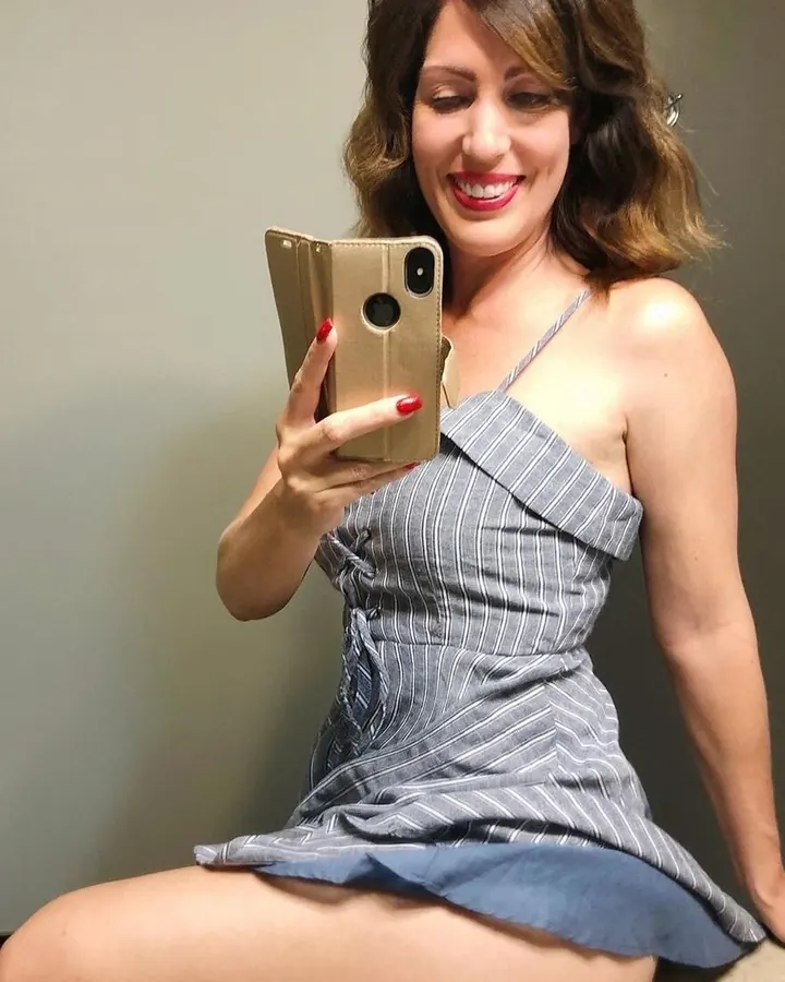 Femme en robe grise à rayures, prenant un selfie avec un téléphone doré, assise sur un canapé.