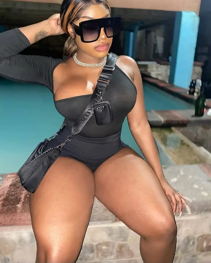 Femme en combinaison noire, lunettes de soleil, sac à bandoulière, assise près d'une piscine, main dans les cheveux.