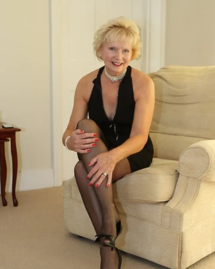 Femme blonde en robe noire, assise sur un canapé beige, souriant à l'appareil, jambes croisées.