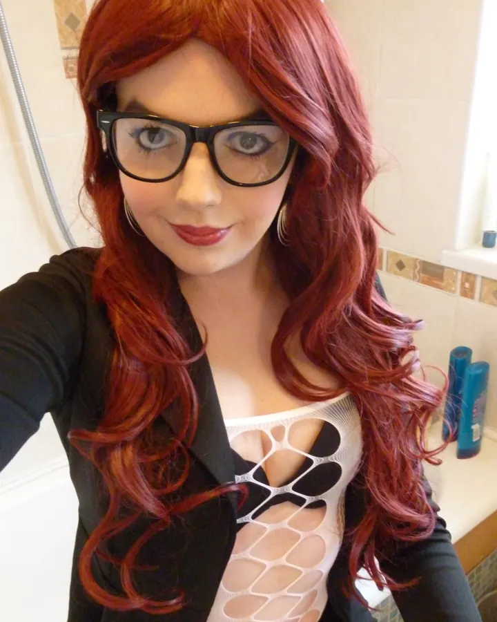 Femme aux cheveux roux, lunettes noires, blouse noire, body blanc ajouré, prenant un selfie dans une salle de bain.