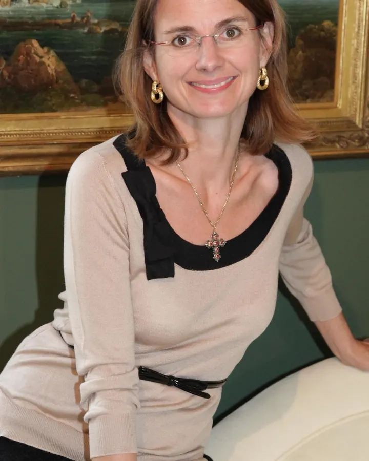 Femme aux cheveux châtains, lunettes fines, collier croix, souriant à l'appareil devant un tableau.