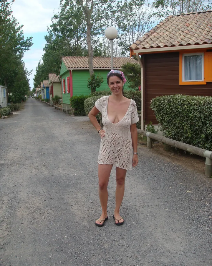 Femme souriante en robe blanche ajourée, debout sur un chemin de camping entre des chalets colorés