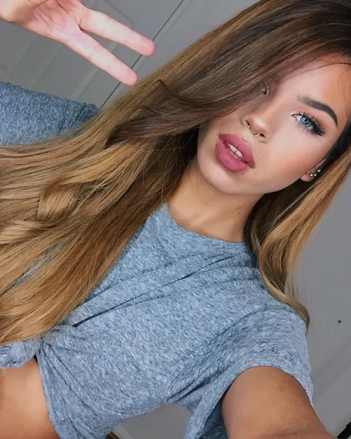 Jeune femme aux longs cheveux bruns clairs en t-shirt gris, faisant un signe de victoire devant l'objectif