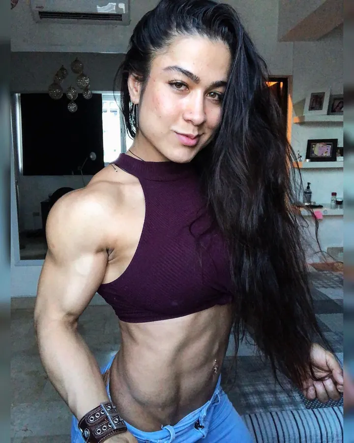 Femme aux cheveux bruns-noirs en haut violet et jeans, montrant ses muscles, regardant l'appareil photo.
