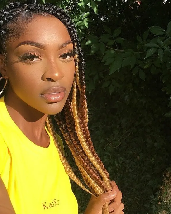 Femme aux cheveux bruns-noirs en t-shirt jaune, tenant une mèche de cheveux, regardant l'appareil photo, avec feuillage en arrière-plan.