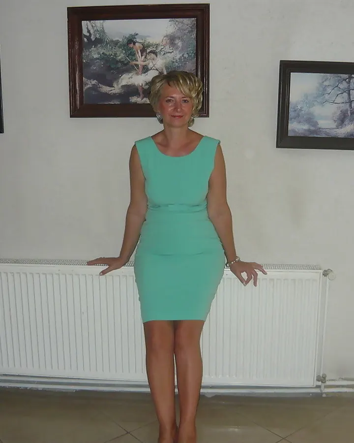 Femme blonde en robe turquoise, debout devant un radiateur, souriante, regardant l'appareil photo.