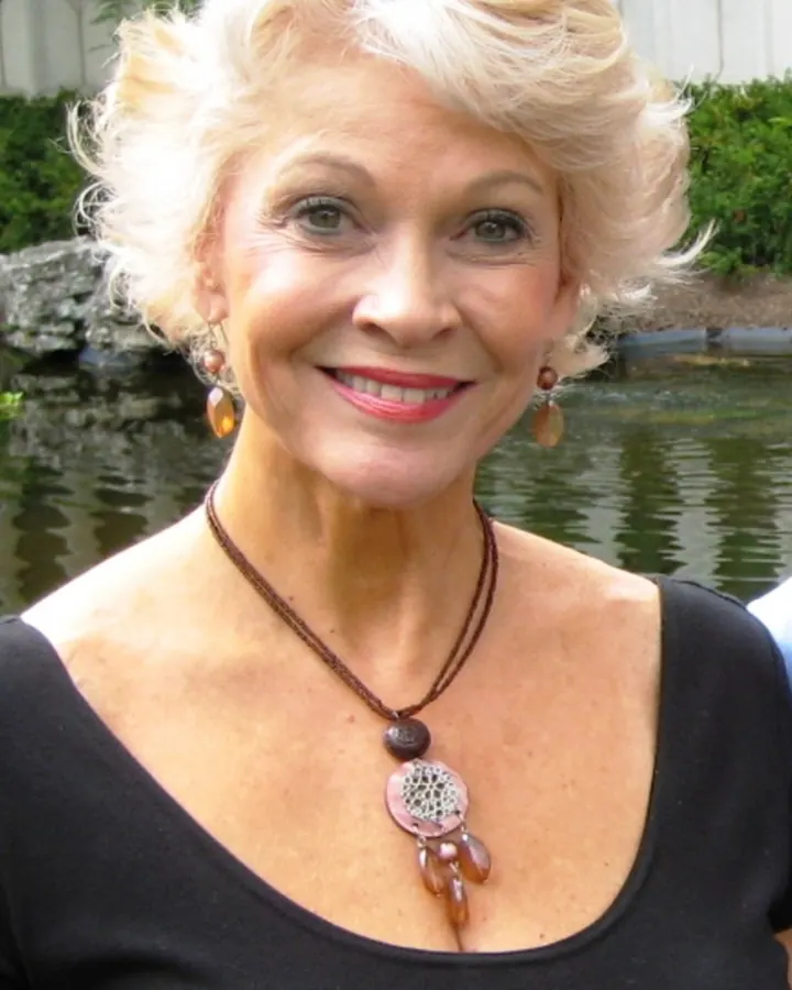 Femme aux cheveux blancs courts, souriante, portant un collier et des boucles d'oreilles, en extérieur près d'un étang