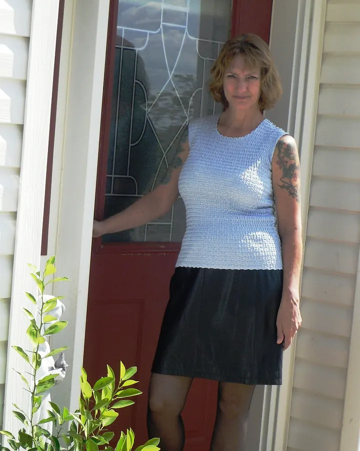 Femme en haut bleu clair et jupe noire, tatouages aux bras, debout devant une porte rouge