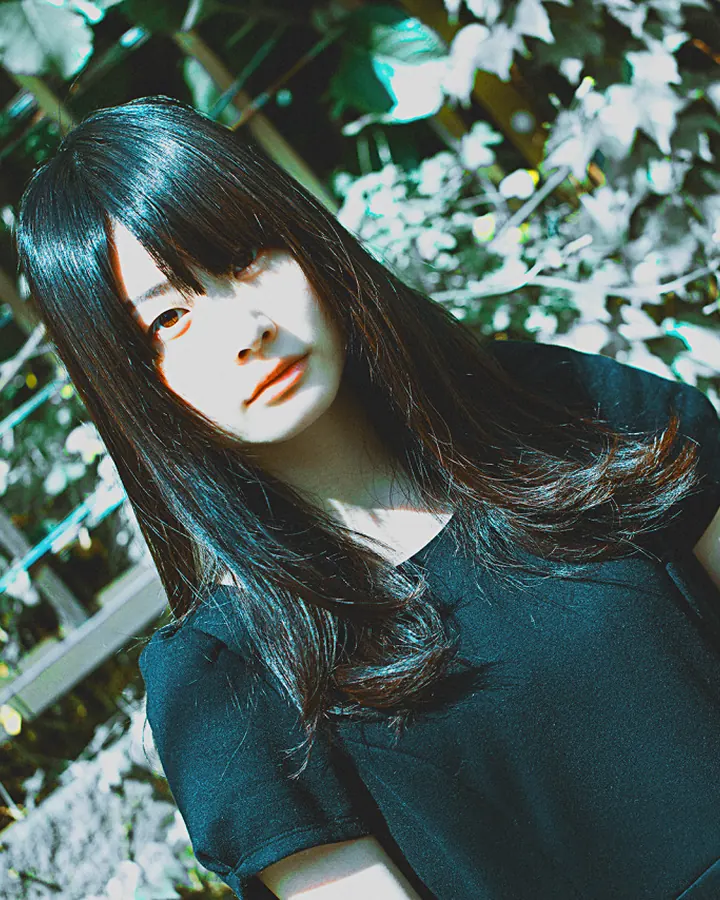 Jeune femme asiatique aux cheveux noirs, en robe noire, posant dans un jardin verdoyant avec un effet de lumière bleu-vert.