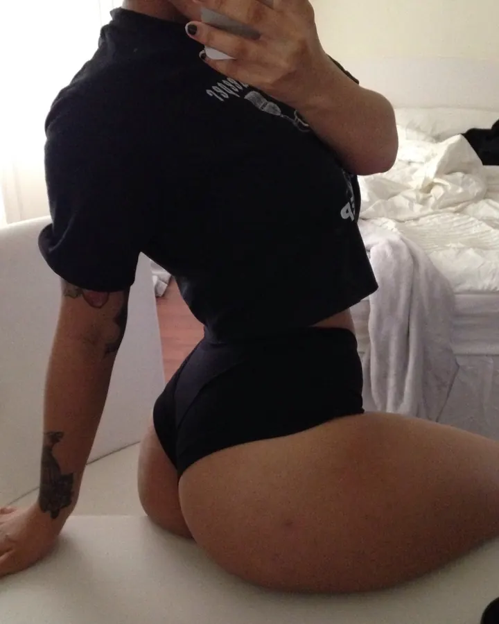 Femme en tenue noire, vue de dos, assise sur une table blanche, tenant un téléphone, avec un tatouage visible sur le bras