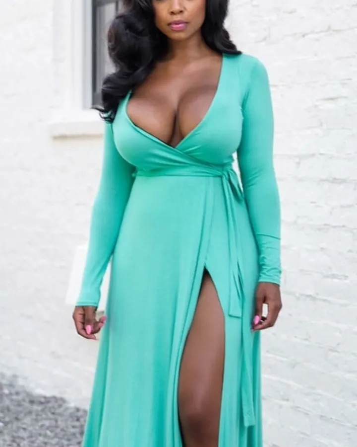 Femme aux cheveux noirs portant une robe verte longue, posant devant un mur blanc, avec une fente haute.