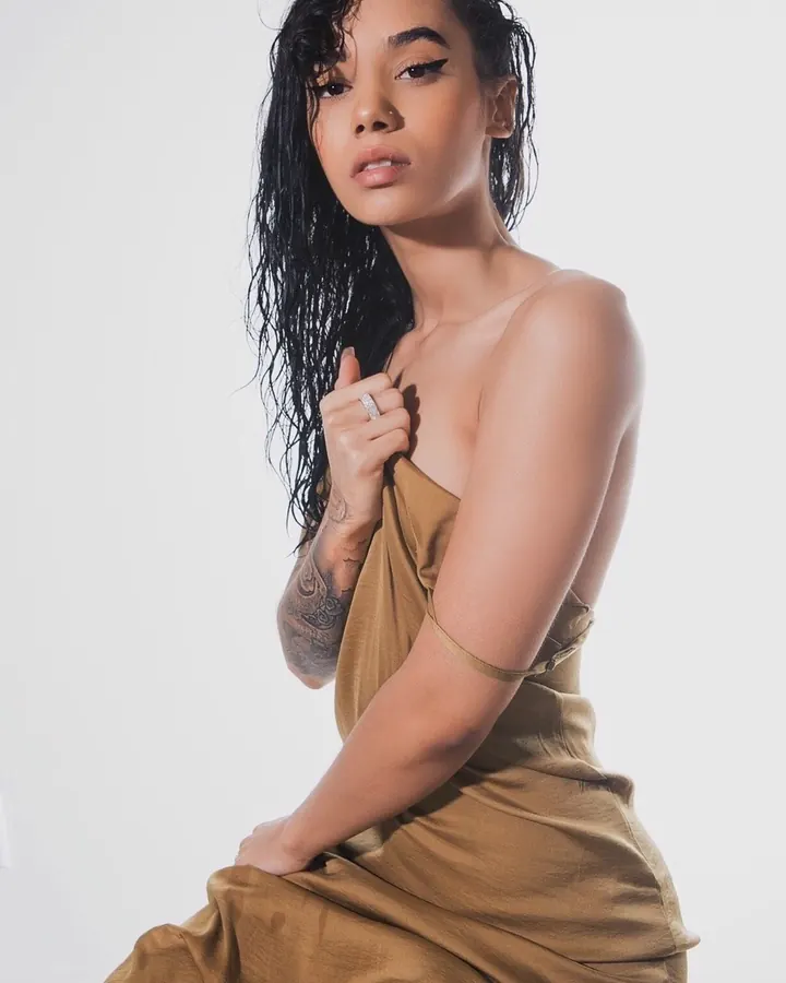 Femme aux cheveux noirs portant une robe marron, assise, tenant son collier, avec des tatouages visibles sur le bras.