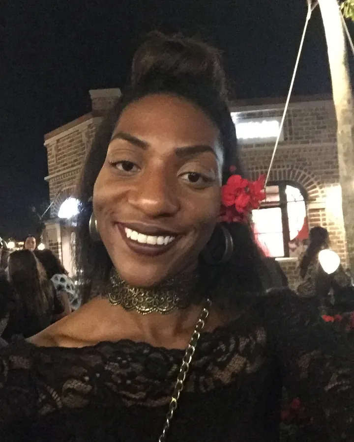 Femme aux cheveux noirs portant un haut noir, posant de profil contre un mur blanc, avec un tatouage visible sur le bras.