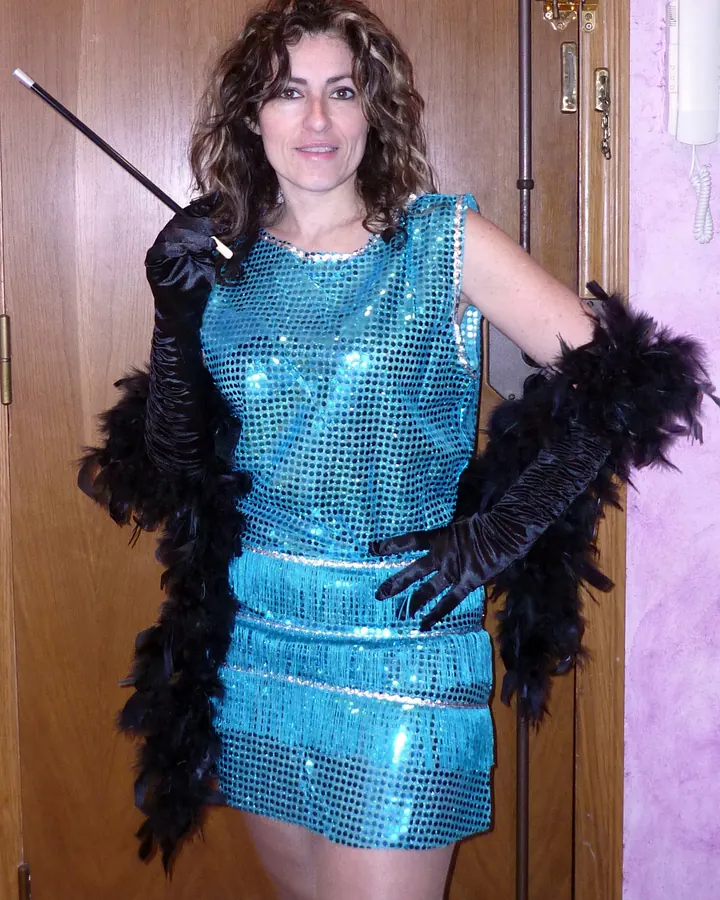 Femme aux cheveux bruns bouclés, portant une robe courte turquoise scintillante, des gants noirs ornés de plumes et tenant une canne, devant une porte en bois.