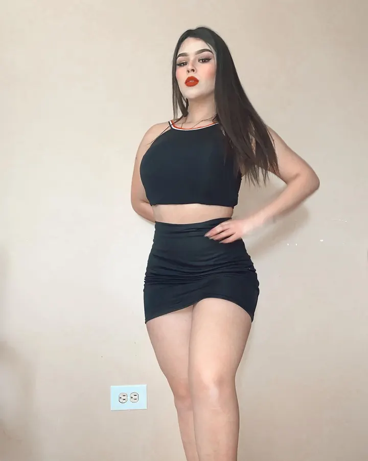 Jeune femme aux cheveux longs noirs, vêtue d'un haut noir et d'une jupe courte assortie, posant contre un mur beige avec une prise électrique visible.