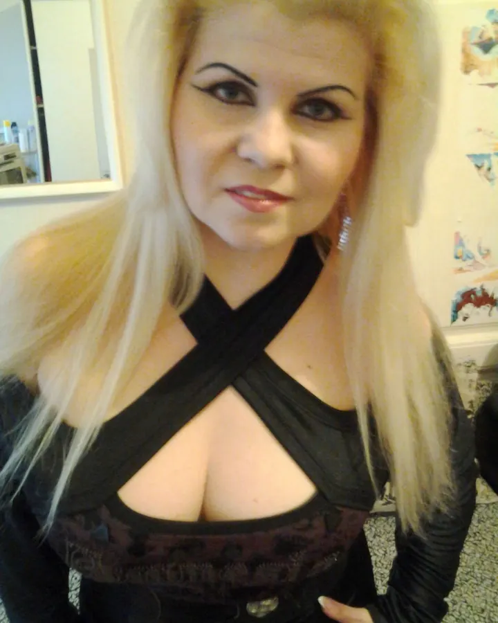 Femme aux cheveux blonds longs, portant un haut noir à découpes croisées, avec des boucles d'oreilles pendantes, dans un cadre intérieur.