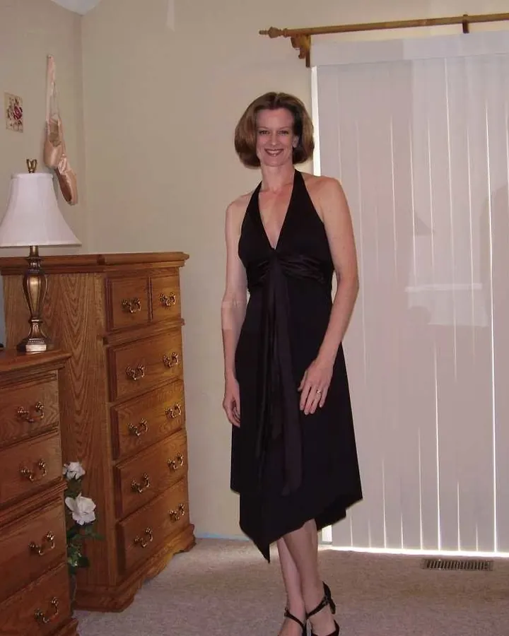 Femme blonde aux yeux bleus, souriante, portant une robe noire, debout dans une chambre avec un grand meuble en bois et un miroir.